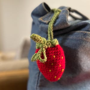 Puede incluir: Una fresa roja de ganchillo con hojas verdes colgando de una bolsa de mezclilla.