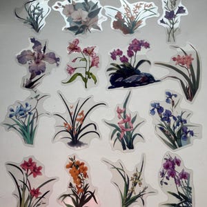 May include: A selection of floral stickers with watercolour flower designs. The stickers depict various blooms in shades of purple, pink, orange, and blue, with green foliage. The stickers are cut out and ready to use.