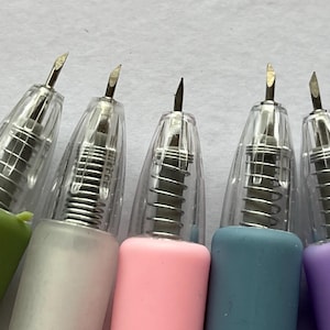 May include: A close-up of five craft knives with angled blades. The knives have clear plastic housings, silver springs, and colourful handles in green, white, pink, blue, and purple. The blades are silver and angled.
