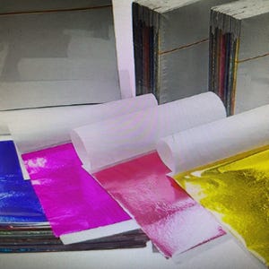 May include: A selection of craft materials, featuring stacks of paper, rolls of white paper, and sheets of metallic foil in blue, pink, and gold. The items are arranged, suggesting a crafting or art project.