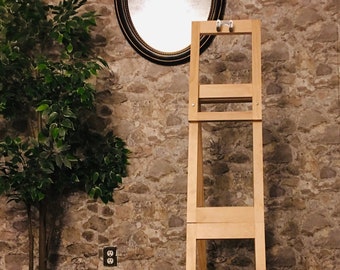 May include: A tall, unfinished wooden plant stand with a rectangular frame. The stand has a simple design with horizontal supports and two metal hooks at the top. The background features a stone wall, a mirror, and a potted plant.