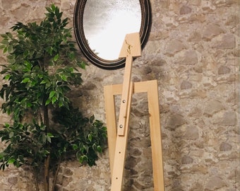 May include: A wooden easel with a light brown finish. The easel is made of two long pieces of wood that are connected by a metal hinge. The easel is standing on a dark wood floor.