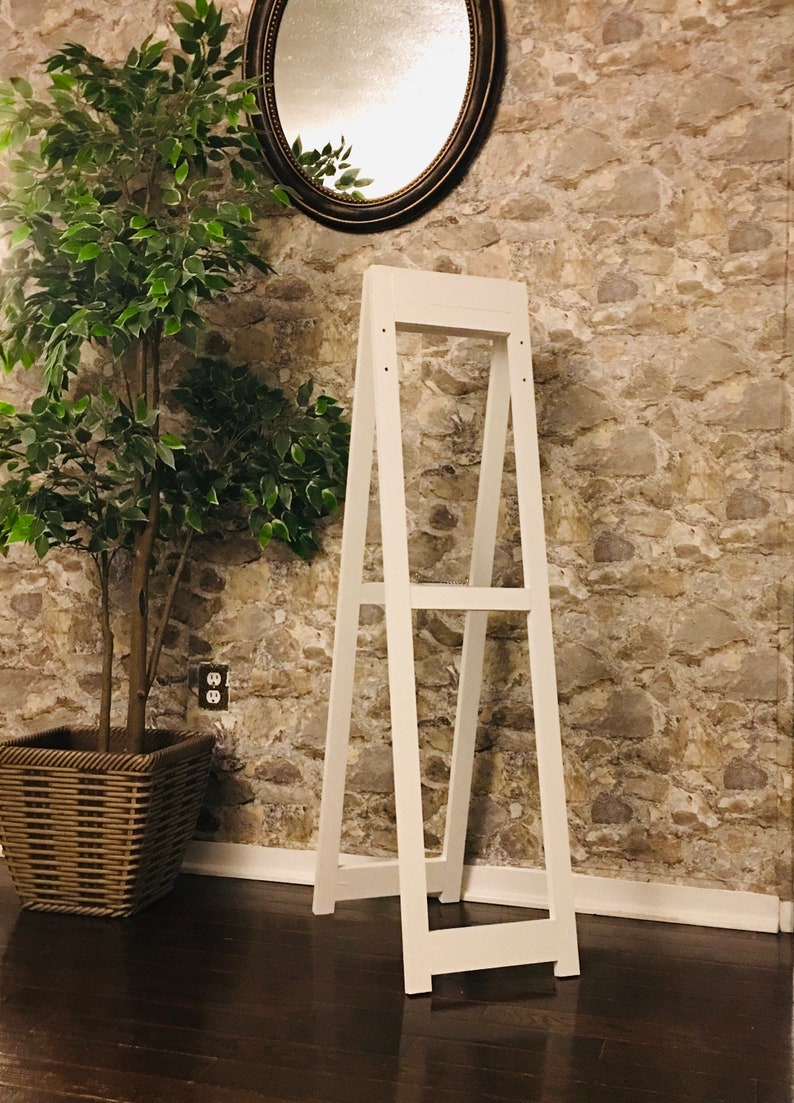 May include: White wooden A-frame display stand with two shelves. The stand is in front of a stone wall with a mirror above it.