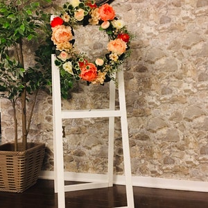May include: A white wooden easel with a floral wreath made of peach, pink, and white flowers. The wreath is hanging on the easel and is in front of a stone wall.