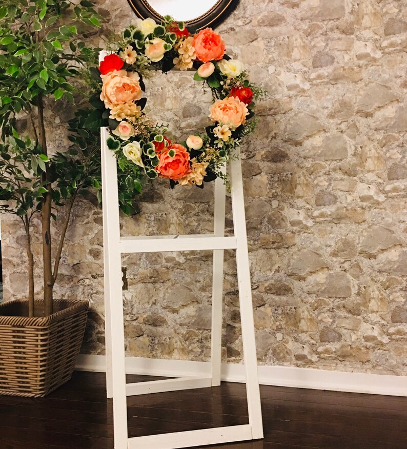 May include: A white wooden easel with a floral wreath made of peach, pink, and white flowers. The wreath is hanging on the easel and is in front of a stone wall.