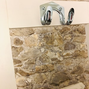 May include: A silver metal hook with two prongs mounted on a white wall with a stone wall background.