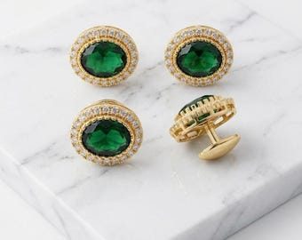 May include: Four gold cufflinks with oval emerald-green stones and a halo of small, clear gemstones. One cufflink is shown on its side, displaying the gold backing and post. The cufflinks are displayed on a white marble surface.