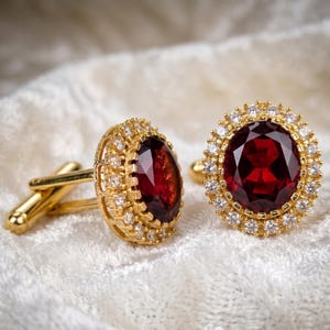 Puede incluir: Un par de gemelos de oro con piedras de granate rojo oscuro de corte ovalado como piezas centrales. Cada granate está rodeado por un halo de pequeñas piedras blancas de corte redondo engastadas en oro, con un mecanismo de cierre de oro pulido.