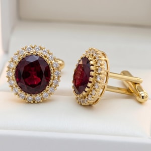 May include: A pair of gold-toned cufflinks with a large, round, deep red gemstone center, surrounded by a halo of small, clear crystals. The cufflinks are presented in a white box, showcasing their elegant design.