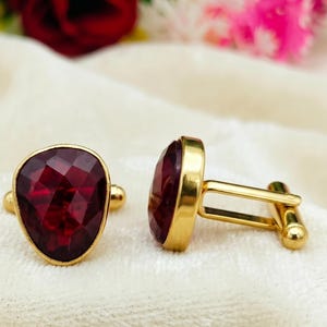 Può includere: Una coppia di gemelli color oro con pietre rosso scuro a forma di pera sfaccettate. Un gemello è posizionato frontalmente per mostrare il taglio geometrico, mentre l'altro è girato per rivelare il profilo laterale e la chiusura metallica a perno.