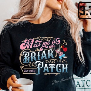 Pode incluir: Camisola de cor preta com um design gráfico rosa e azul que diz "Meet me at the Briar Patch Est. 1989". O gráfico inclui um coelho de desenho animado a segurar um saco.