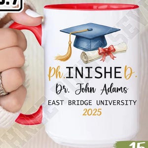 May include: White 15oz ceramic mug with a red interior and handle. The mug features a graduation cap, diploma, and the text "ph.INISHED Dr. John Adams EAST BRIDGE UNIVERSITY 2025".