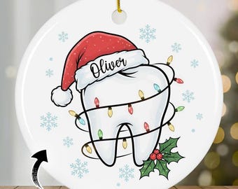 May include: A white ceramic Christmas ornament featuring a cartoon tooth wearing a Santa hat with the name "Oliver". The tooth is wrapped in colorful Christmas lights and decorated with snowflakes and holly.