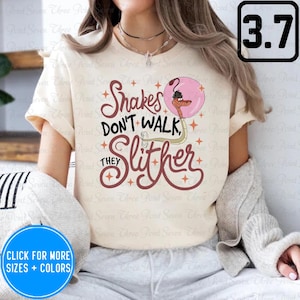 May include: A white t-shirt with a pink and brown graphic that says "Snakes Don't Walk, They Slither". The graphic also includes a cartoon snake in a pink balloon.