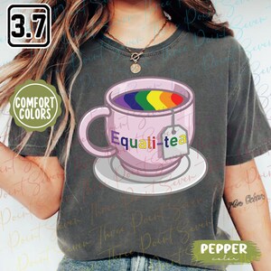 Può includere: T-shirt grigia con una tazza rosa contenente tè arcobaleno e la scritta "Equali-tea". La tazza è su un piattino bianco, con una bustina di tè. La maglietta ha anche il testo "Comfort Colors" e "Pepper color".