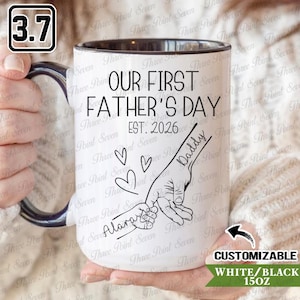 May include: White ceramic mug with a black rim and handle. The mug features the text "OUR FIRST FATHER'S DAY EST. 2026" and a graphic of two hands holding each other. The word "Daddy" is written on one hand and "Always" on the other.