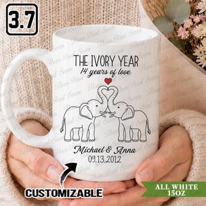 May include: A white ceramic mug featuring a black line drawing of two elephants with trunks intertwined to form a heart shape. Above the elephants, the text reads THE IVORY YEAR 14 years of love. Below, the names Michael & Anna and the date 13.09.2012 are printed.