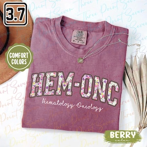May include: Berry-coloured t-shirt featuring floral "HEM-ONC" lettering and "Hematology Oncology" text. The shirt has a crew neck and a small Comfort Colors tag. A silver necklace with a pendant is draped on the shirt.