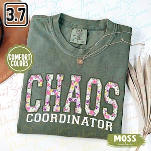 May include: A green t-shirt with the text "CHAOS COORDINATOR" in white letters surrounded by pink, purple, and yellow flowers. The shirt is made by Comfort Colors and is in the color moss.