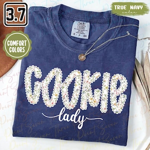 Könnte beinhalten: Ein marineblaues Comfort Colors T-Shirt mit den Worten "COOKIE lady" in einem floralen Design. Das Shirt hat einen Rundhalsausschnitt und kurze Ärmel. Eine goldene Halskette mit Anhänger ist ebenfalls sichtbar.