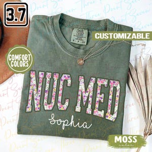 May include: A green t-shirt with the text "NUC MED" in white letters outlined in purple. The letters are filled with a floral pattern. The text "Sophia" is written below the letters in white. The text "COMFORT COLORS" is on a white tag attached to the shirt.