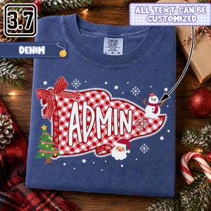 May include: A denim blue crew neck t-shirt featuring a red and white gingham pennant graphic with the text ADMIN in white. The design includes a red bow, a green Christmas tree, a snowman, a Santa Claus face, and white snowflakes.