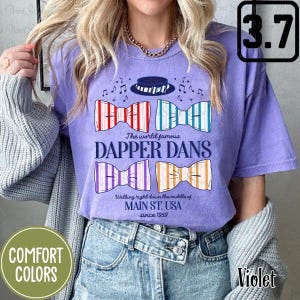 Op de afbeelding: Een paars T-shirt met een afbeelding van vier strikjes en de tekst "The world famous Dapper Dans Walking right down the middle of MAIN ST. USA since 1959".