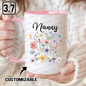 May include: White ceramic mug with a pink handle and interior, featuring the word "Nanny" in black script. The mug is adorned with a colorful floral design, including various flowers and butterflies. The mug is customizable.