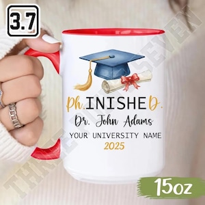 May include: White ceramic mug with a red interior and handle. The mug features a graduation cap, diploma, and the words "ph.INISHED" and "Dr. John Adams" with "YOUR UNIVERSITY NAME 2025" printed on it. The mug holds 425g.