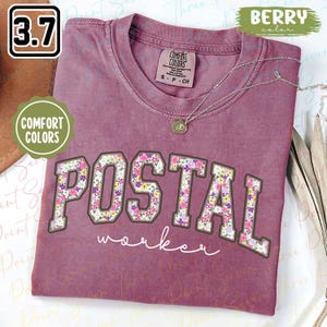 Könnte beinhalten: Beerenfarbenes T-Shirt mit dem Wort "POSTAL" in einem floralen Design und dem Wort "worker" in weißer Schrift. Das Shirt hat einen Rundhalsausschnitt und ein Comfort Colors-Etikett.