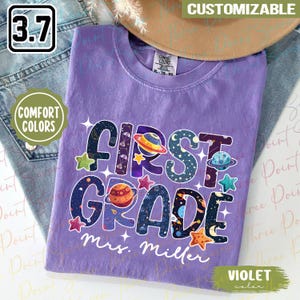 May include: A violet Comfort Colors t-shirt with the words "FIRST GRADE" in a space-themed design. The shirt includes the name "Mrs. Miller" and is customizable. The shirt is folded on top of a pair of jeans.