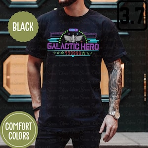 Op de afbeelding: Zwart T-shirt met een grafisch ontwerp van een ruimteschip met de tekst "GALACTIC HERO" en "LEVEL 7" in neonkleuren. De tekst "999999" is ook in het ontwerp opgenomen.