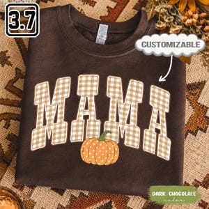May include: A dark chocolate brown crewneck sweatshirt featuring the word MAMA in a beige and white gingham check pattern. Below the text is an orange pumpkin graphic with white polka dots. The sweatshirt includes a tag at the neck.