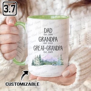 Peut inclure: Mug en céramique blanche avec une anse et un bord intérieur verts. La tasse présente les mots "DAD Est. 1998", "GRANDPA Est. 2020" et "GREAT-GRANDPA Est. 2025". Une scène de montagne et d'arbres à l'aquarelle est en dessous. Le mot "CUSTOMIZABLE" est également visible.