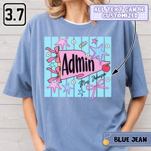 Op de afbeelding: Een blauw t-shirt met een grafische print van een roze wimpel met de tekst "Admin" en "Mrs. Johnson". Het ontwerp bevat roze en blauwe sterren, kleine witte bloemen, een paperclip, een rode appel en roze linten op een lichtblauwe gestreepte achtergrond.