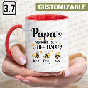 May include: White ceramic mug with a red interior and handle. The mug features the text "Papa's reasons to BEE HAPPY" with bee illustrations and names. The mug is held by a person.