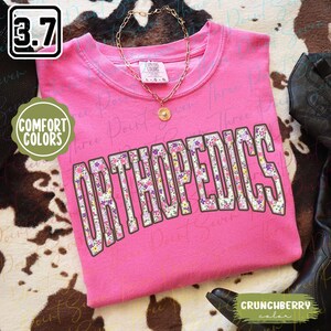 May include: Pink t-shirt with the word "ORTHOPEDICS" in white letters outlined in brown. The letters are filled with a floral pattern in shades of pink, purple, and yellow. The shirt is made of a soft, comfortable fabric.