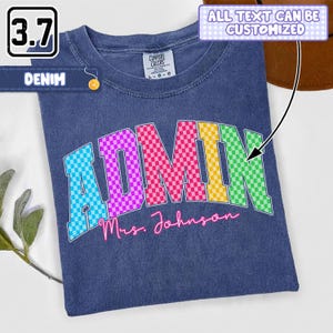 May include: A denim-colored t-shirt featuring the word ADMIN in a multi-colored checkered pattern with the text Mrs. Johnson written in pink script below. The shirt includes a Comfort Colors label at the neck and a note stating all text can be customized.