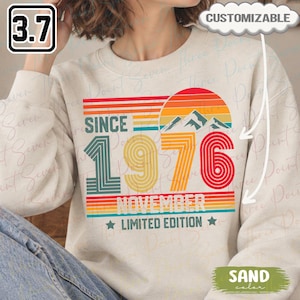 May include: A sand-colored crewneck sweatshirt featuring a retro-style graphic with a sunset, mountains, and the text "SINCE 1976 NOVEMBER LIMITED EDITION". The design uses horizontal stripes in orange, yellow, and teal, with the year 1976 in large, bold, multi-line font.