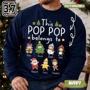 May include: A navy blue crewneck sweatshirt featuring the white text This POP POP belongs to, surrounded by Christmas-themed illustrations of seven children dressed as a gift, tree, snowman, penguin, Santa, star, and elf, with names printed below each.