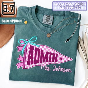 Op de afbeelding: Een blauw-sparrenkleurig t-shirt met een afbeelding van een roze en wit geblokte wimpel met de tekst "ADMIN" in grote donkerpaarse letters en "Mrs. Johnson" in roze cursief schrift. Een lichtblauw lint is aan de wimpel bevestigd.