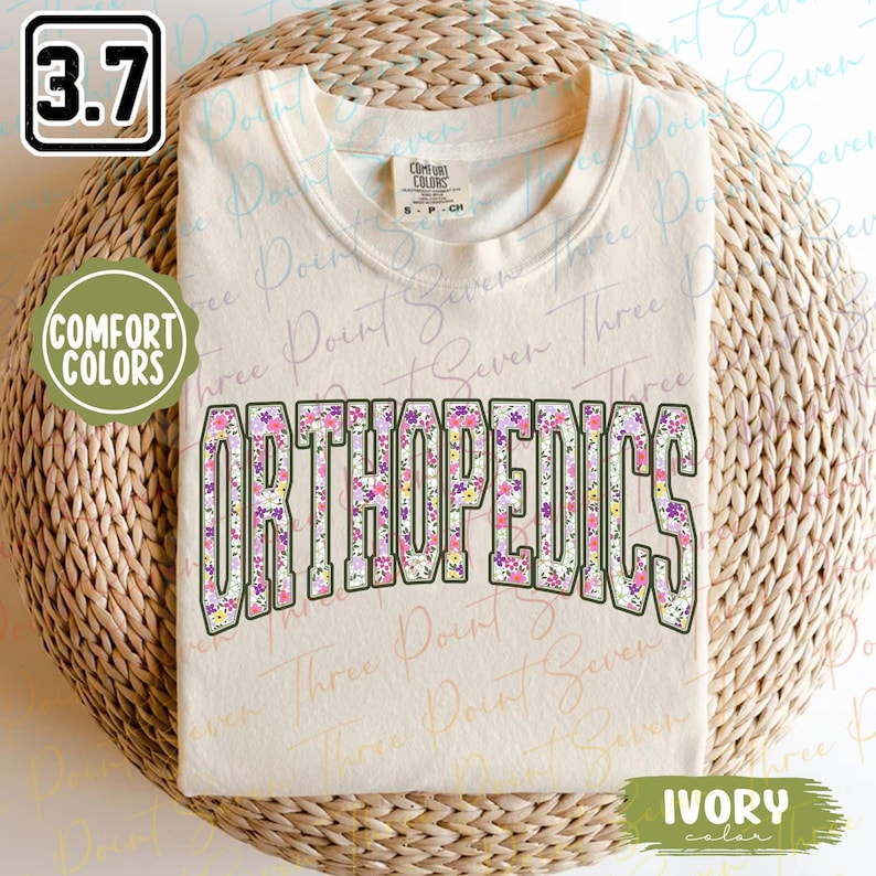 May include: A beige t-shirt with the word "ORTHOPEDICS" printed in white letters outlined in black. The letters are filled with a floral pattern in shades of pink, purple, and yellow. The shirt is labeled "COMFORT COLORS" and "IVORY color".