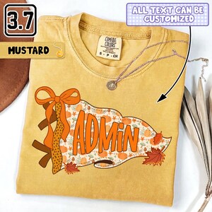 May include: A mustard yellow t-shirt featuring a graphic of a pennant flag with the text ADMIN in orange letters. The flag design includes a pattern of pumpkins and leaves, accented by a large orange bow and a leopard print ribbon tail.
