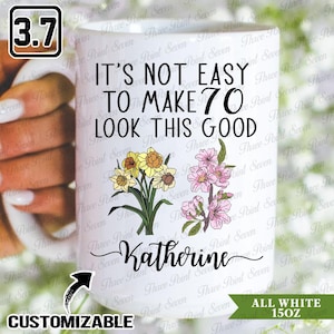 Peut inclure: Mug blanc personnalisable de 15 oz (environ 425 ml) avec le texte "IT'S NOT EASY TO MAKE 70 LOOK THIS GOOD" et le nom "Katherine". Il est orné d'illustrations florales de jonquilles et de fleurs roses.