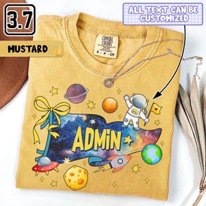 Op de afbeelding: Een mosterdgeel t-shirt met een afbeelding van een ruimte-thema wimpel met de tekst ADMIN, omringd door planeten, een maan, een raket, sterren en een astronaut die een vlag vasthoudt. Het shirt heeft een ronde hals en een Comfort Colors label.