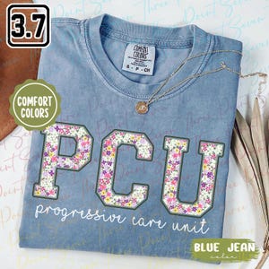 May include: Blue jean colored t-shirt with the letters "PCU" in a floral pattern. The words "progressive care unit" are below the letters. A gold necklace with a pendant is on the shirt.
