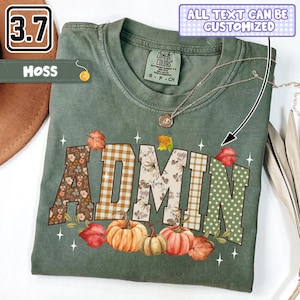 May include: A moss green t-shirt featuring the word ADMIN in large block letters with varied patterns including floral, plaid, and polka dots. The design is accented with orange and green pumpkins, autumn leaves, and small star shapes.