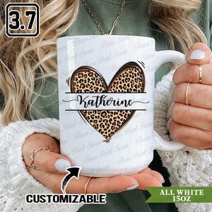 Puede incluir: Taza de cerámica blanca de 15 oz con un diseño de corazón con estampado de leopardo. El corazón está dividido por una línea horizontal con el nombre "Katherine" en cursiva. La taza es personalizable.