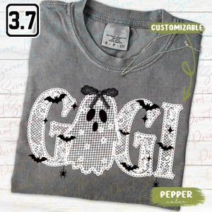 May include: A charcoal gray t-shirt featuring a white lace-patterned graphic that spells GAGI, with the letter A replaced by a ghost wearing a black bow. The design includes small black bats and spiders, with text reading 3.7 and CUSTOMIZABLE.