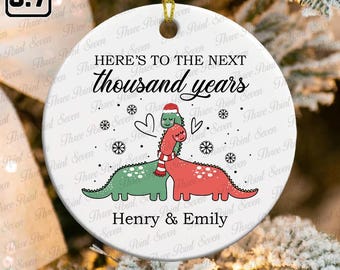 May include: A white ceramic ornament with a gold string for hanging. The ornament features a green and red dinosaur couple hugging, with text that reads "HERE'S TO THE NEXT thousand years" and names "Henry & Emily".